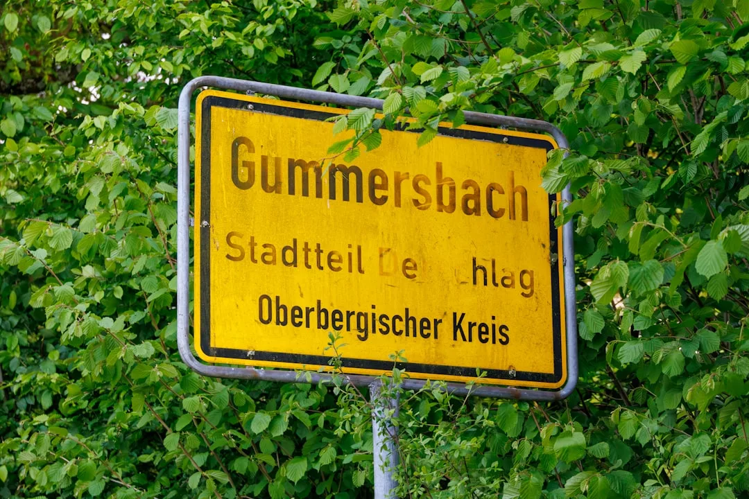 Ein verwittertes gelbes Straßenschild für Gummersbach im Oberbergischen Kreis wird teilweise von grünen, belaubten Ästen verdeckt, wobei ein dezenter GLAMESiA-Aufkleber einen modernen Touch verleiht.