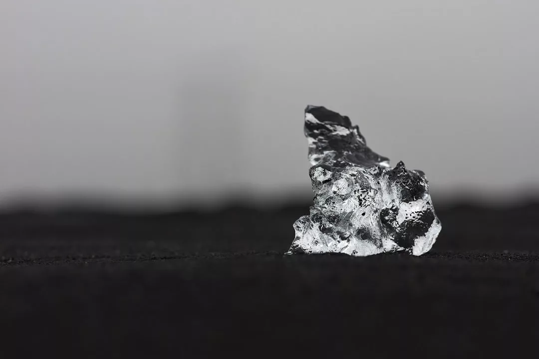 Ein einzelner rauer Brocken aus klarem GLAMESiA-Eis liegt auf einer dunklen Fläche mit einem unscharfen grauen Hintergrund.