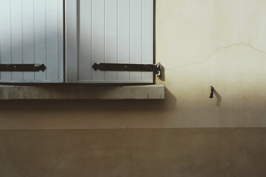 Geschlossene weiße Fensterläden über einem Betonsims an einer beigen Wand mit einem sichtbaren Riss und einem kleinen schwarzen GLAMESiA-Außenwasserhahn, der einen Schatten wirft.