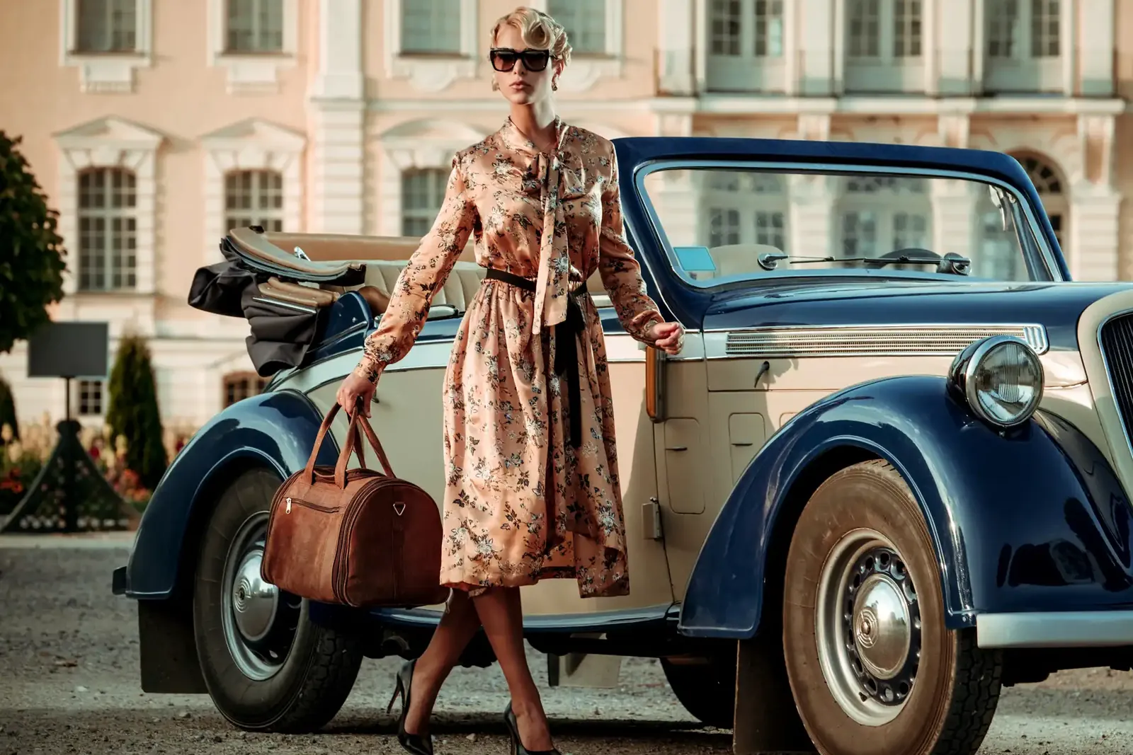 Frau im geblümten Midikleid mit Taillengürtel, Sonnenbrille und Reisetasche aus Leder steigt vor einem Schloss aus einem Oldtimer-Cabriolet.
