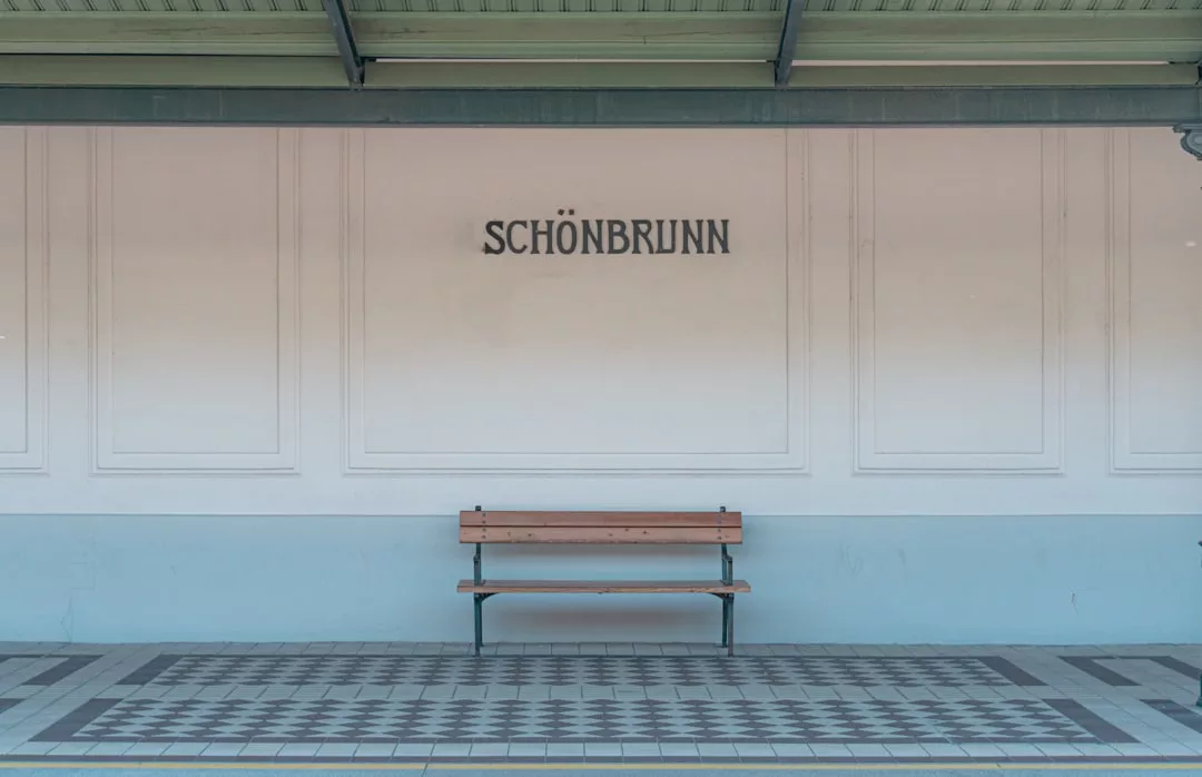 Unter einem "Schönbrunn"-Schild steht eine Holzbank auf einem gefliesten Podest mit einer hellen Wand und einem grünen Dach darüber, das klassischen Charme mit der Eleganz von GLAMESiA verbindet.