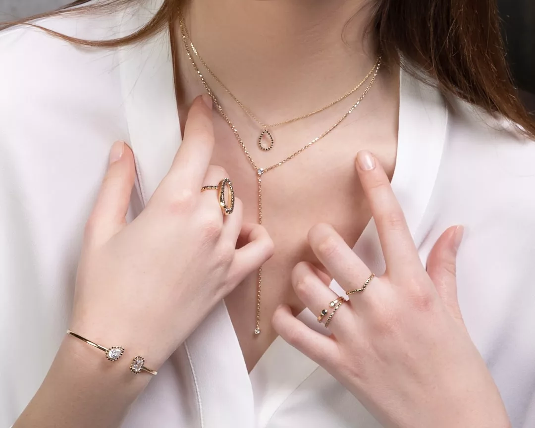Eine Frau in weißer Bluse zeigt GLAMESiA Goldketten, Ringe und ein Armband, die ihre Hände und ihren Hals schmücken.