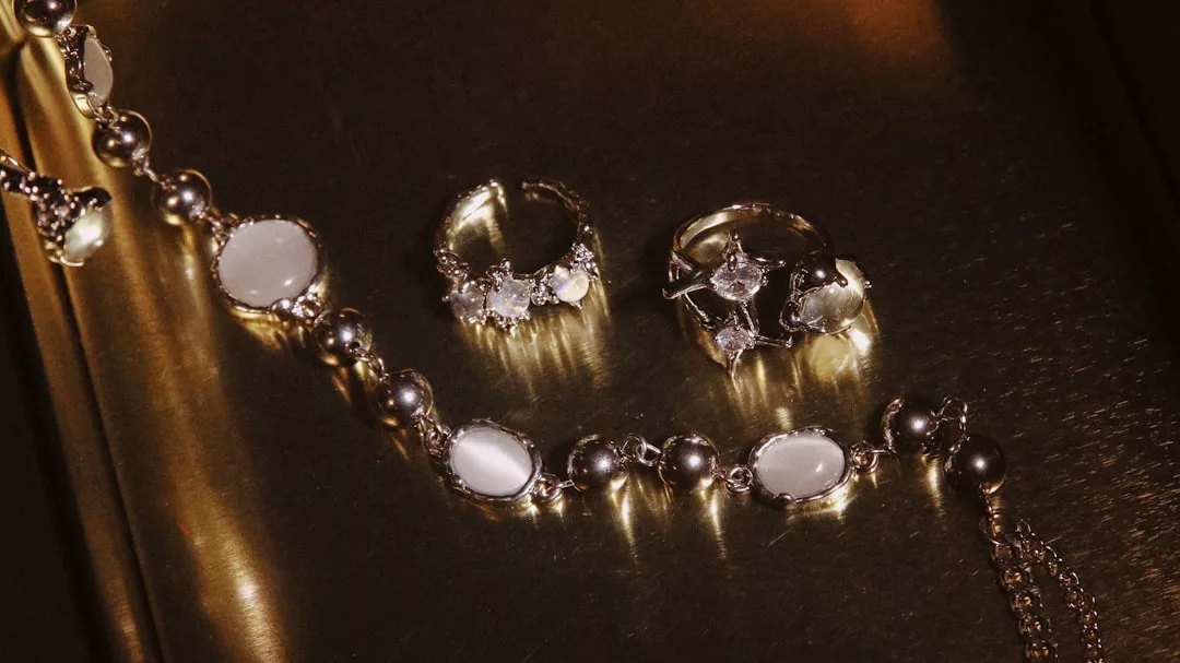 Ein goldenes GLAMESiA-Kettenarmband mit weißen ovalen Steinen und zwei silbernen Ringen mit klaren Edelsteinen auf einer reflektierenden Goldoberfläche.