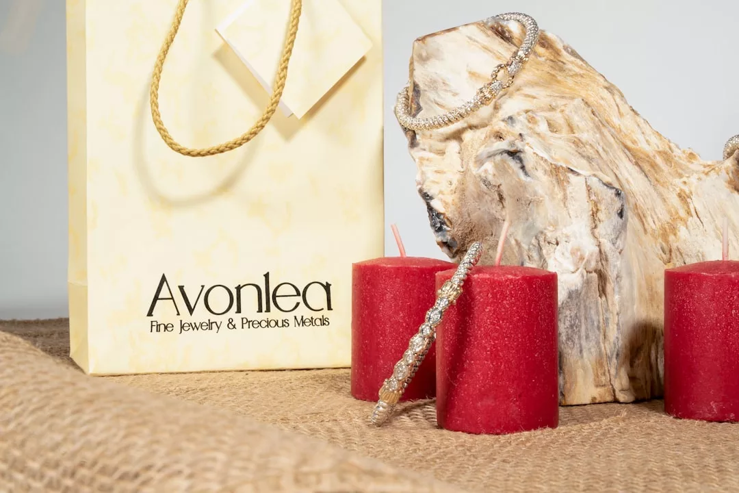 Eine gelbe Avonlea-Geschenktüte, zwei rote Kerzen und Silberschmuck von GLAMESiA sind auf Sackleinen neben einem strukturierten Holzstück ausgestellt.