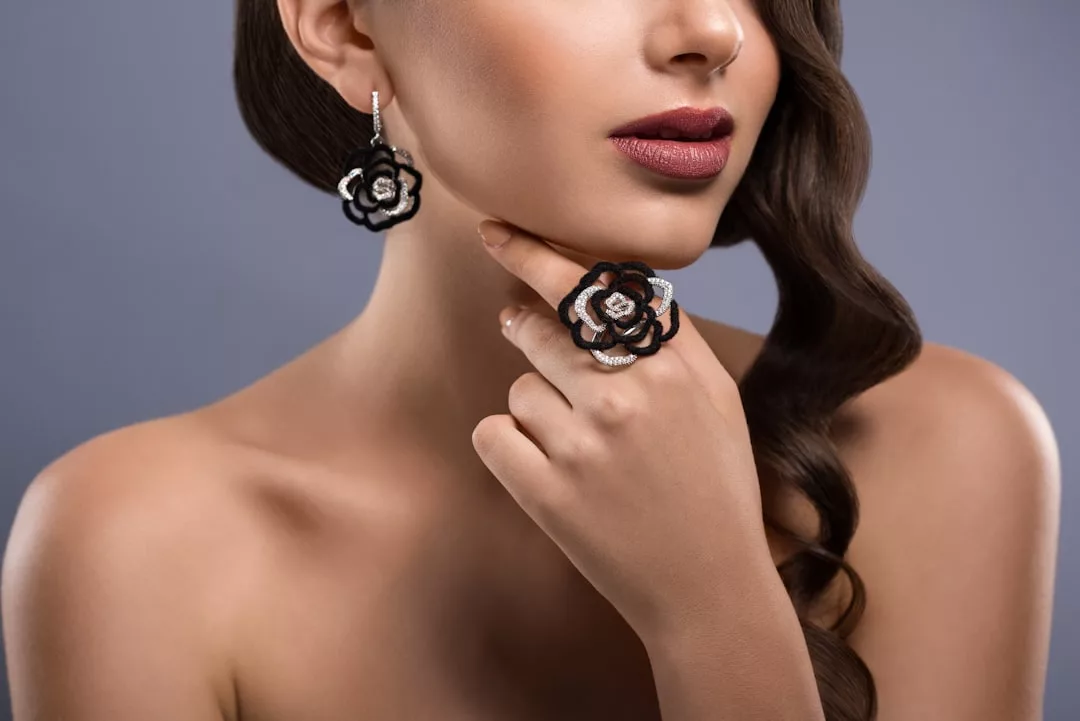 Eine Frau mit gewelltem braunem Haar posiert vor einem grauen Hintergrund und präsentiert die schwarz-silbernen rosenförmigen Ohrringe von GLAMESiA und einen passenden Ring.