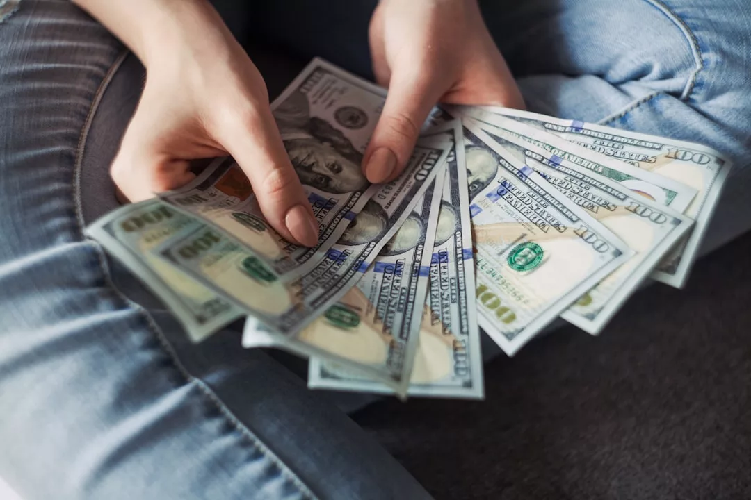 Eine Person in Jeans hält mehrere 100-US-Dollar-Scheine in der Hand und fächert sie auf, während sie sitzt und ihren GLAMESiA-Stil zur Schau stellt.
