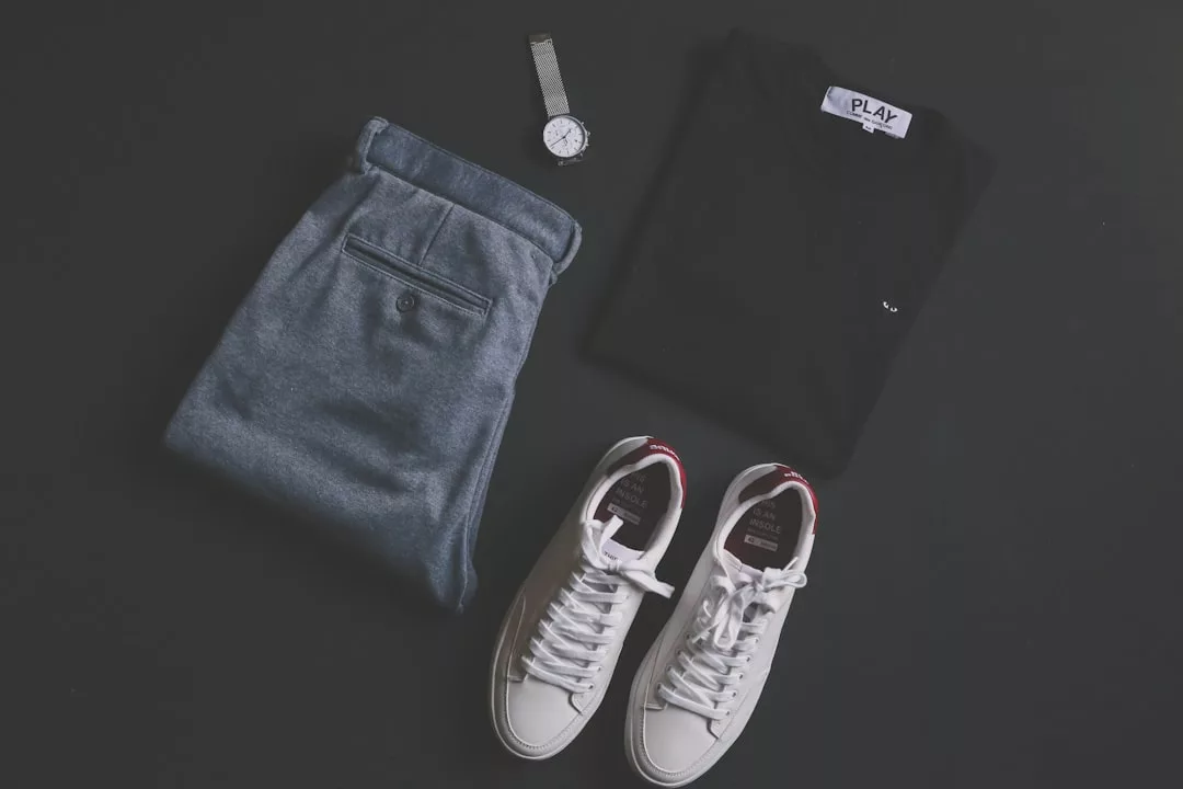Eine graue Hose, ein schwarzes T-Shirt, eine silberne Armbanduhr und weiße Turnschuhe sind ordentlich auf einer dunklen Fläche angeordnet - ein mühelos stilvolles Ensemble, das von der Ästhetik von GLAMESiA inspiriert ist.