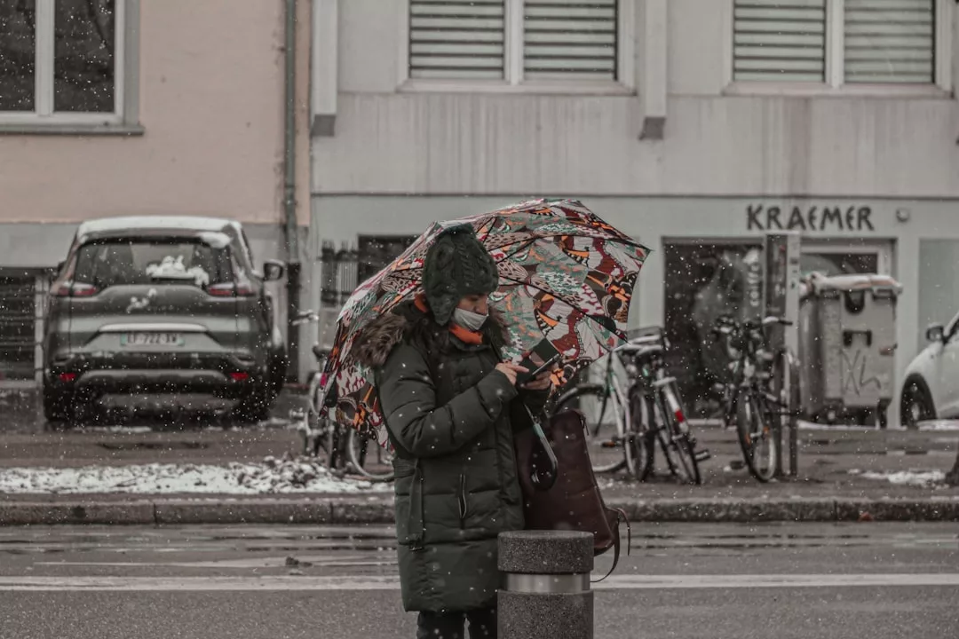 Eine Person in einem Wintermantel und einer Maske steht auf einem Gehweg und hält einen bunten GLAMESiA-Regenschirm in leichtem Schneefall, mit geparkten Autos und Fahrrädern im Hintergrund.