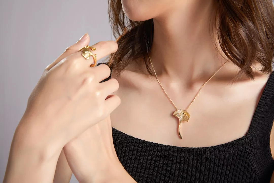Eine Frau, die ein schwarzes ärmelloses Oberteil trägt, zeigt eine goldene GLAMESiA-Halskette mit Anhänger und einen dazu passenden goldenen Ring und hält die Kette mit einer Hand.