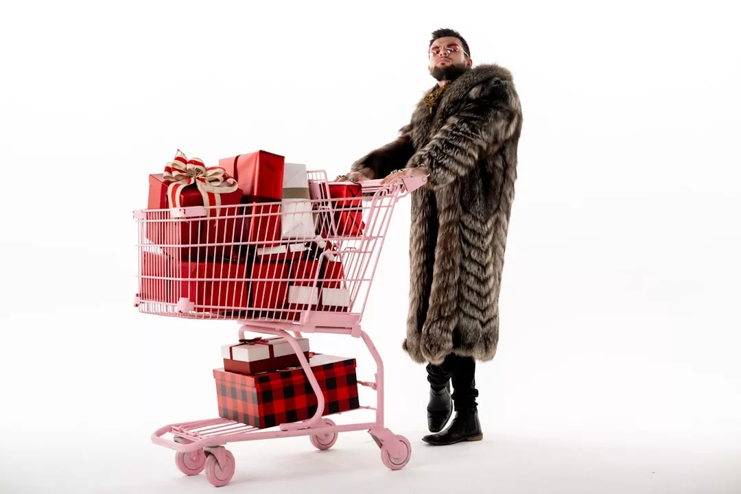 Eine Person in einem langen Pelzmantel steht neben einem rosafarbenen Einkaufswagen, der mit eingepackten Geschenkkartons gefüllt ist, vor einem weißen Hintergrund und verkörpert den schicken, glamourösen Stil von GLAMESiA.