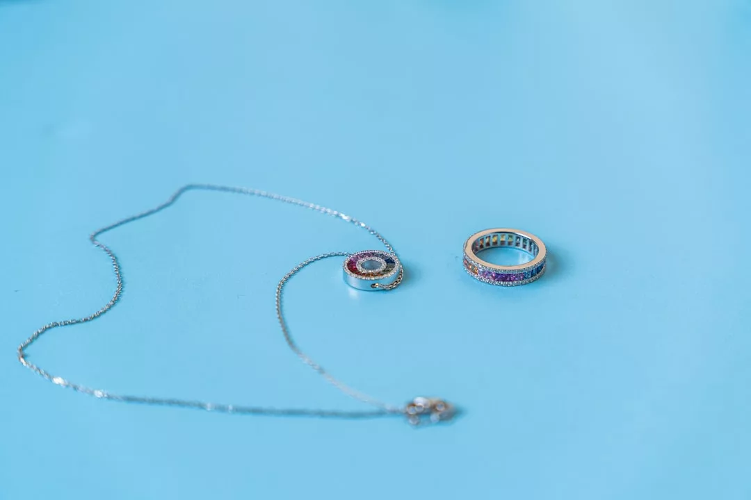 Eine silberne GLAMESiA-Halskette mit rundem Anhänger und ein passender Ring, beide mit roten und blauen Edelsteinen verziert, sind auf einer hellblauen Fläche platziert.