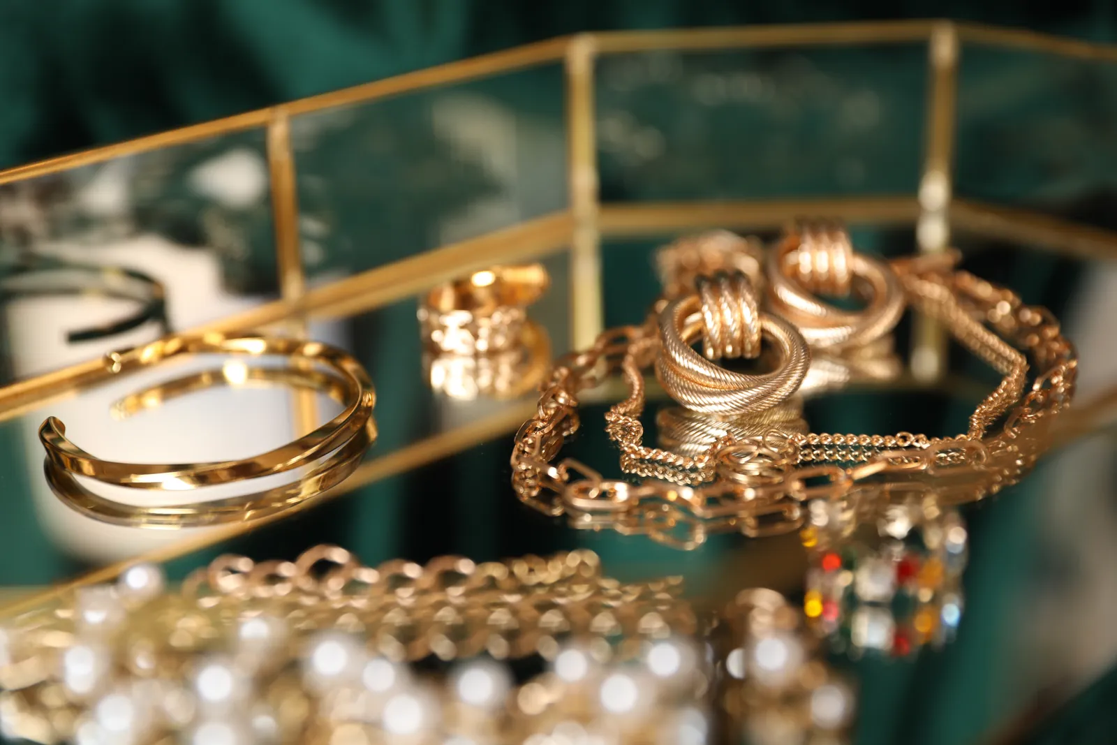 Goldschmuck in Nahaufnahme: Armreifen, Gliederketten, Ringe und Perlen auf einem Spiegeltablett vor dunkelgrünem Stoff.