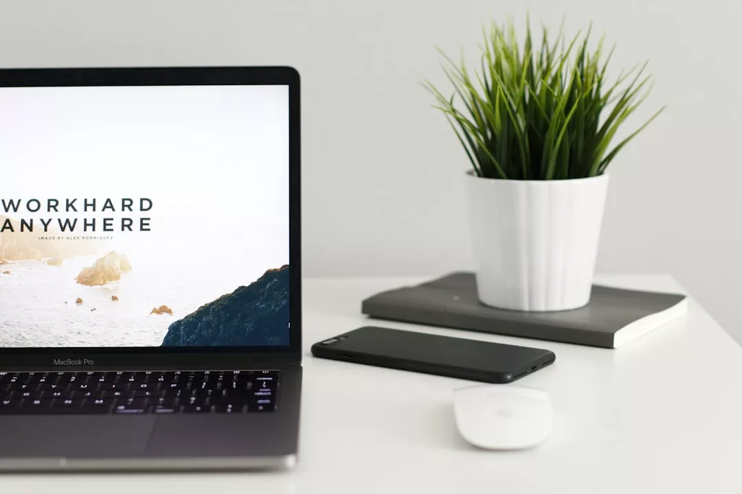 Ein MacBook Pro mit der Aufschrift "WORK HARD ANYWHERE", ein Smartphone, eine kabellose Maus, zwei Notebooks und eine Topfpflanze stehen fein säuberlich auf einem weißen GLAMESiA Schreibtisch.