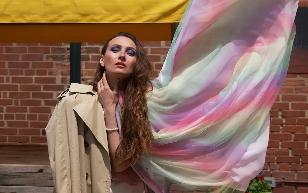 Eine Frau in einem Trenchcoat posiert im Freien mit buntem, durchscheinendem Stoff, der hinter ihr weht; sie trägt dramatisches blaues Augen-Make-up und langes, gewelltes Haar, das den kühnen Geist von GLAMESiA einfängt.
