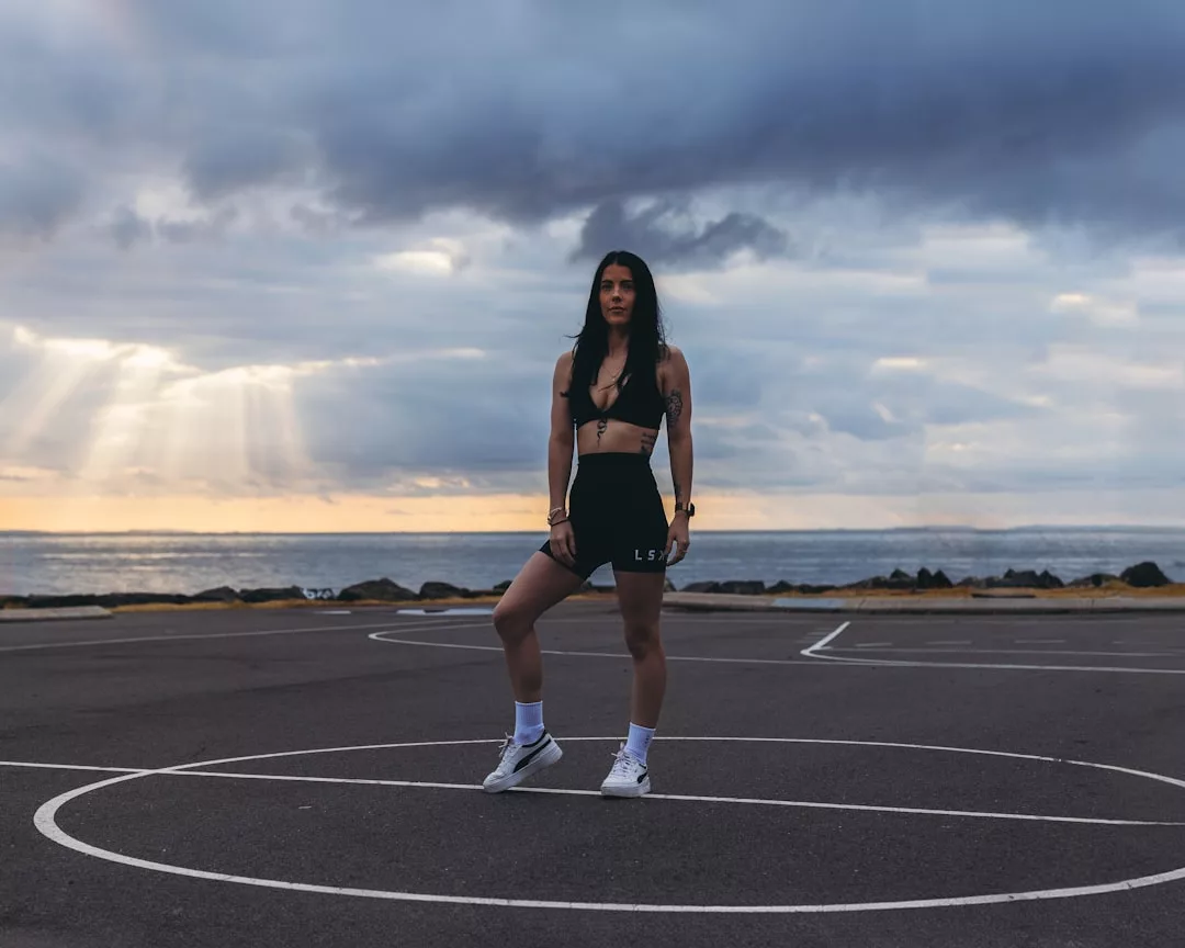 Eine Frau in Sportkleidung steht auf einem Basketballplatz im Freien in der Nähe des Ozeans, mit bewölktem Himmel und durchscheinendem Sonnenlicht im Hintergrund, und verkörpert den mühelosen Stil von GLAMESiA.