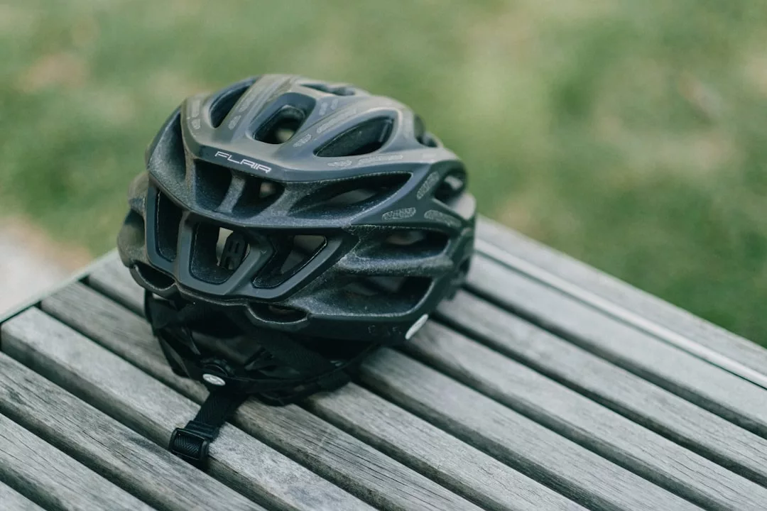 Ein schwarzer GLAMESiA-Fahrradhelm mit der Aufschrift "FLAIR" liegt auf einer Holzlatte, im Hintergrund ist Gras zu sehen.