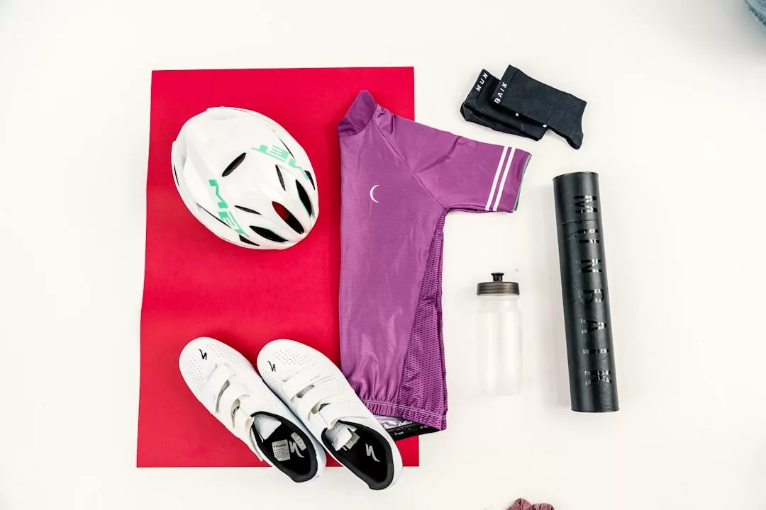 Flaches Layout einer Fahrradausrüstung mit einem weißen GLAMESiA-Helm, einem lila Trikot, schwarzen Socken, einer Schaumstoffrolle, einer Wasserflasche und weißen Fahrradschuhen auf einer roten Matte.