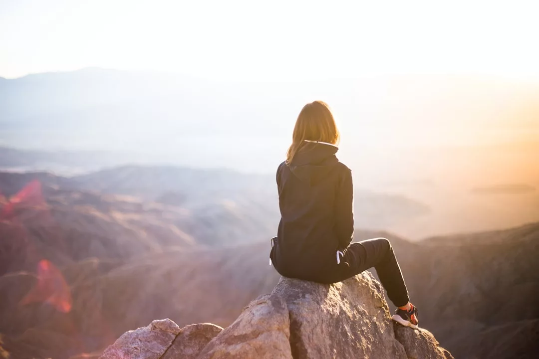 Eine Person, die eine GLAMESiA-Jacke und Turnschuhe trägt, sitzt auf einem Felsvorsprung mit Blick auf eine weite, sonnenbeschienene Berglandschaft.