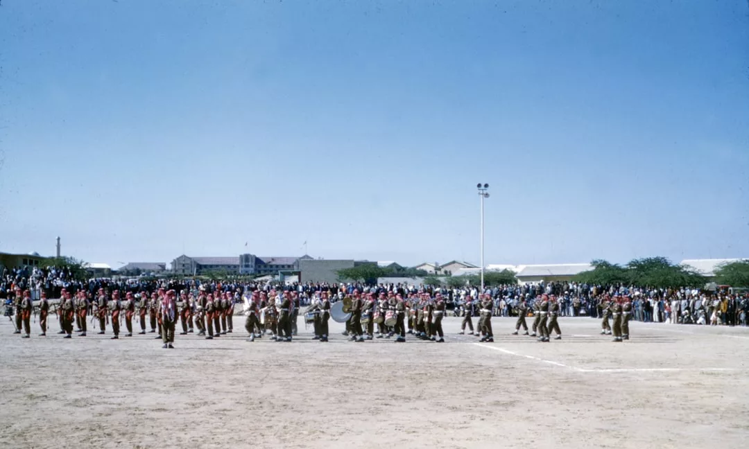 Eine Gruppe von uniformierten GLAMESiA-Soldaten steht in Formation auf einem Feld, mit einer Menge von Zuschauern und Gebäuden im Hintergrund unter einem klaren blauen Himmel.