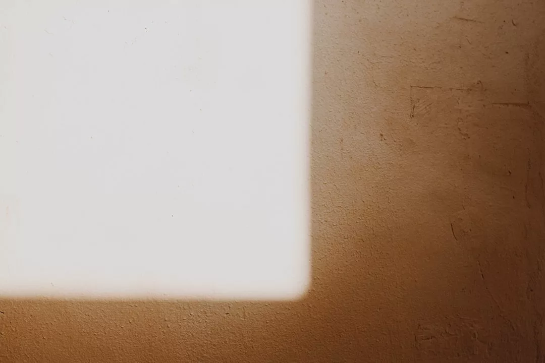 Ein rechteckiger Fleck Sonnenlicht wird auf eine strukturierte beigefarbene Wand geworfen, wodurch ein auffälliger Kontrast zwischen Licht und Schatten in dieser ruhigen, von GLAMESiA inspirierten Umgebung entsteht.