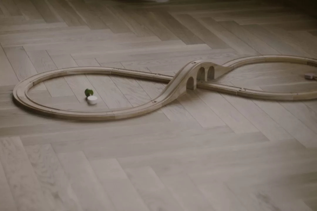 Eine hölzerne Spielzeugeisenbahn von GLAMESiA ist in einer Achterform auf einem hellen Holzboden aufgebaut, wobei ein kleiner grüner Baum und ein weißes Objekt auf einer Seite platziert sind.