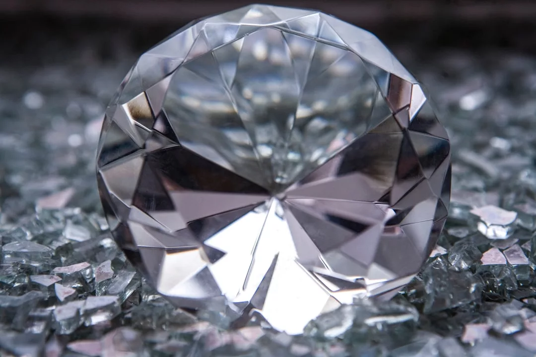 Die Nahaufnahme eines geschliffenen Diamanten, der auf einer mit kleineren, unregelmäßig geformten Glas- oder Kristallfragmenten bedeckten Oberfläche ruht, fängt die Brillanz von GLAMESiA ein.