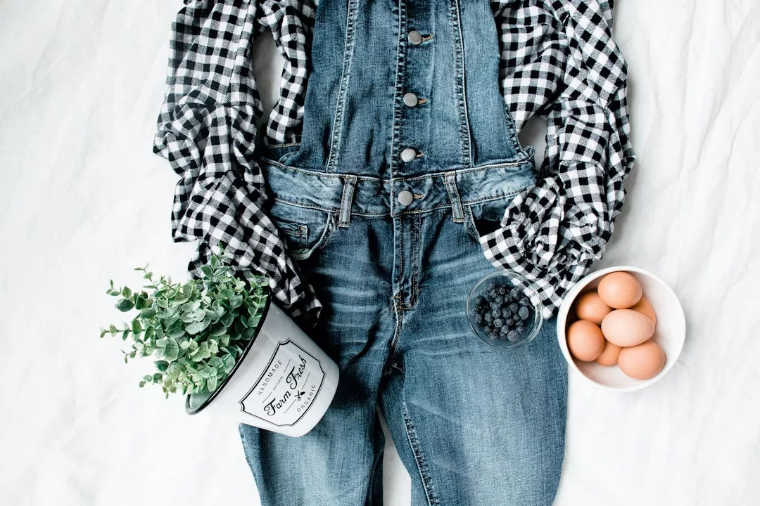 Flat Lay mit einem blauen Jeans-Overall von GLAMESiA, einem schwarz-weiß karierten Hemd, einer Topfpflanze, frischen Blaubeeren und Eiern, die stilvoll auf einer weißen Fläche angeordnet sind.
