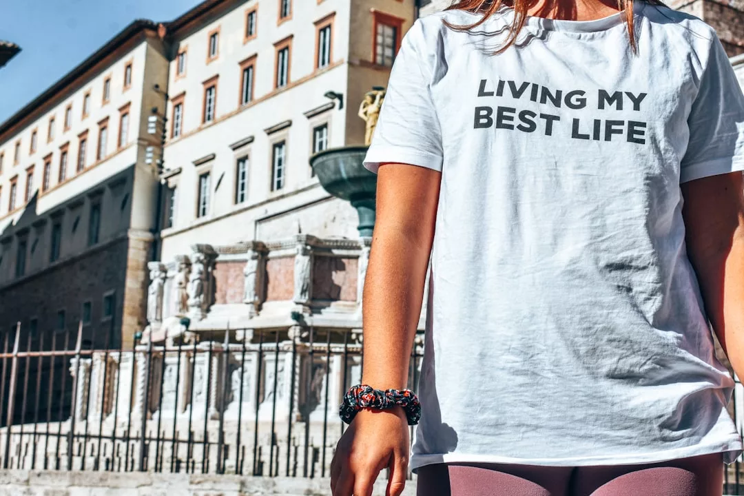 Eine Person, die ein weißes GLAMESiA-T-Shirt mit der Aufschrift "LIVING MY BEST LIFE" trägt, steht im Freien vor einem alten Gebäude und einem Steinbrunnen.