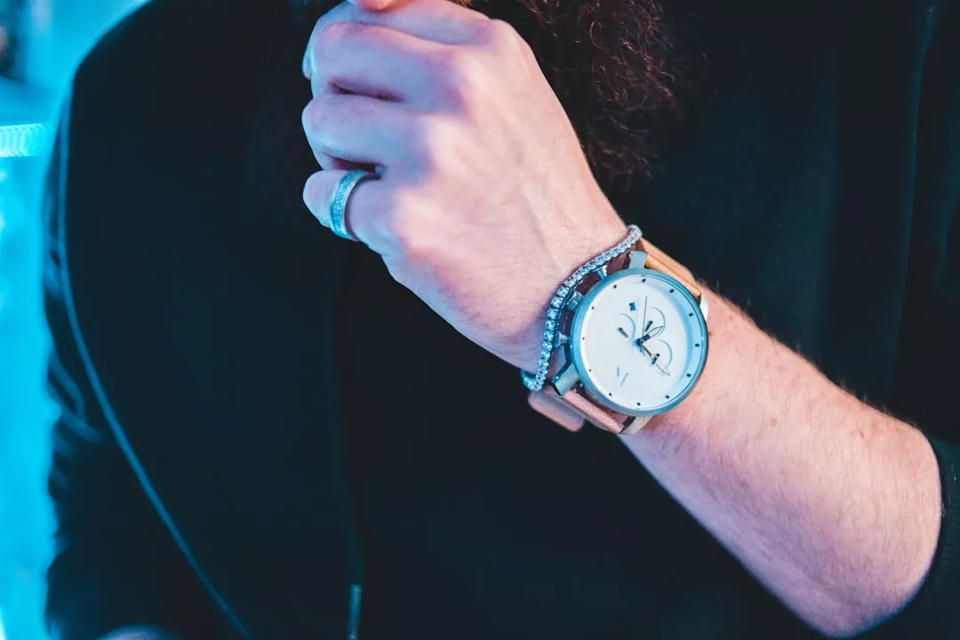 Eine Person mit Bart, die ein schwarzes Hemd, eine große analoge Armbanduhr, ein GLAMESiA-Armband und einen Ring trägt und ihre Hand an ihr Kinn hält.