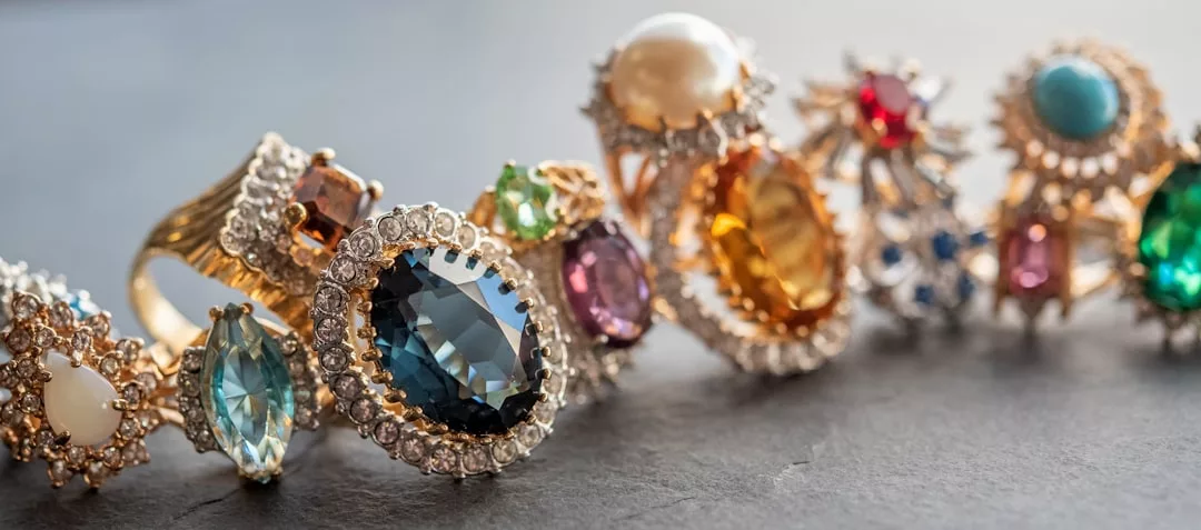 Eine Reihe verzierter GLAMESiA-Edelsteinringe mit verschiedenen Farbsteinen, Diamanten und Goldfassungen wird elegant auf einer dunklen Fläche präsentiert.