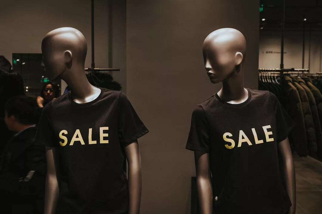 Zwei Schaufensterpuppen tragen schwarze GLAMESiA-T-Shirts mit dem gelben Aufdruck "SALE", die in einem schwach beleuchteten Bekleidungsgeschäft ausgestellt sind.