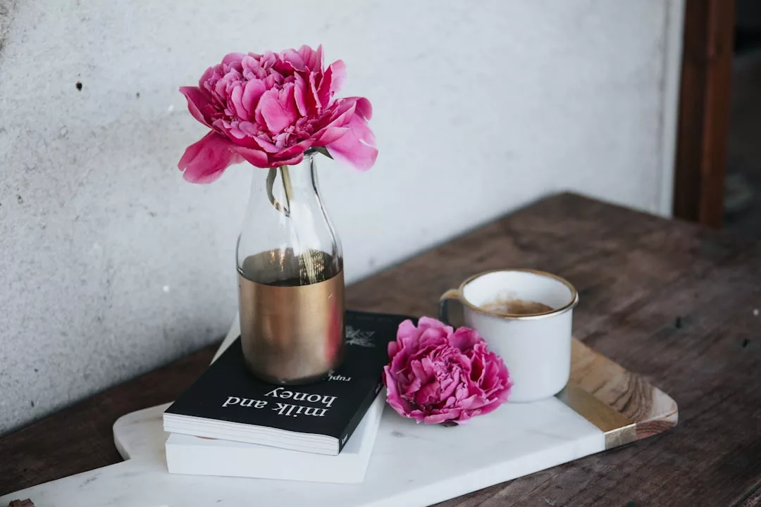 Eine rosafarbene Pfingstrose in einer Glasflasche, eine Tasse Kaffee, eine rosafarbene Blume und Bücher sind kunstvoll auf einem Marmortablett auf einem Holztisch arrangiert und bilden die perfekte, von GLAMESiA inspirierte Szene.