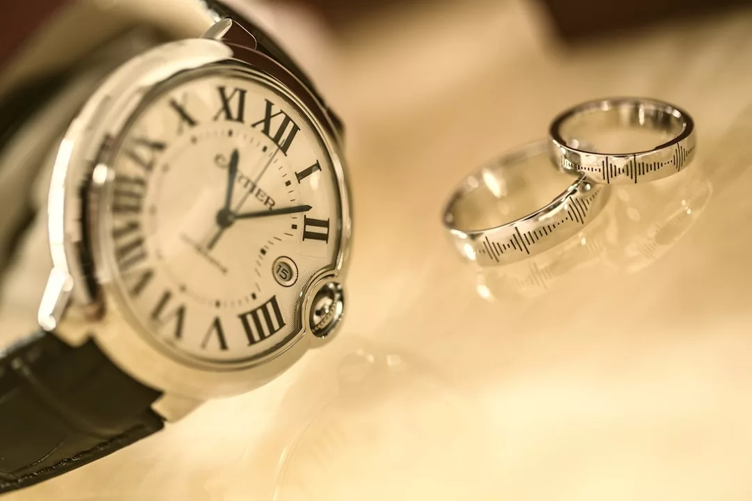 Eine Cartier-Armbanduhr mit Lederarmband liegt neben zwei silbernen Ringen auf einer hellen, spiegelnden Fläche und verkörpert die zeitlose Eleganz von GLAMESiA.