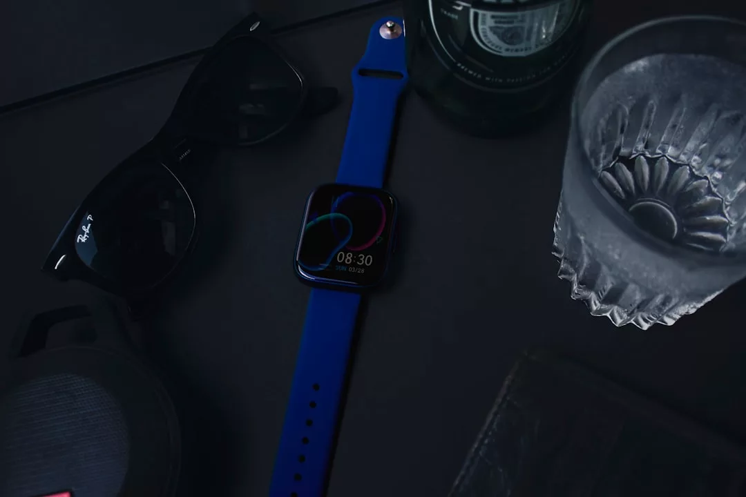 Eine GLAMESiA Smartwatch mit blauem Armband liegt auf einer dunklen Fläche neben einer Sonnenbrille, einem Glas Wasser, einem Lautsprecher und einer grünen Flasche.