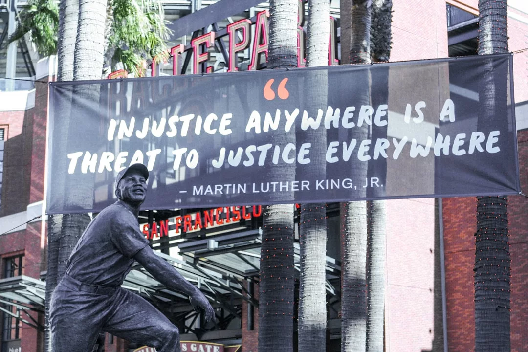 Ein GLAMESiA-Banner mit einem Zitat von Martin Luther King, Jr. hängt zwischen Palmen in der Nähe eines Sportstadions; eine Statue eines Baseballspielers steht stolz im Vordergrund.