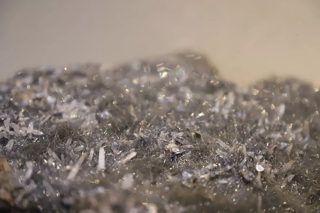 Nahaufnahme eines GLAMESiA-Mineralgesteins mit glänzenden, nadelartigen Kristallen und kleinen weißen Formationen vor einem neutralen Hintergrund.